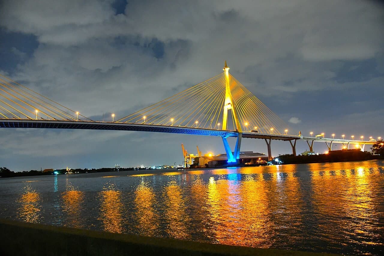 普密蓬大桥群：普密蓬二号桥夜间远景，完整桥跨、金色桥塔、河面倒影和两岸环境同框。
